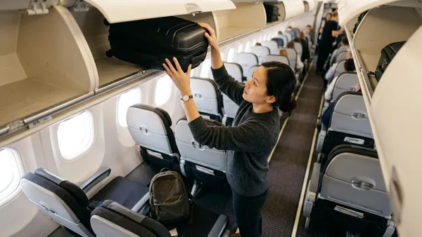 Passenger stowing cabin bag in British Airways overhead locker with underseat backpack visible