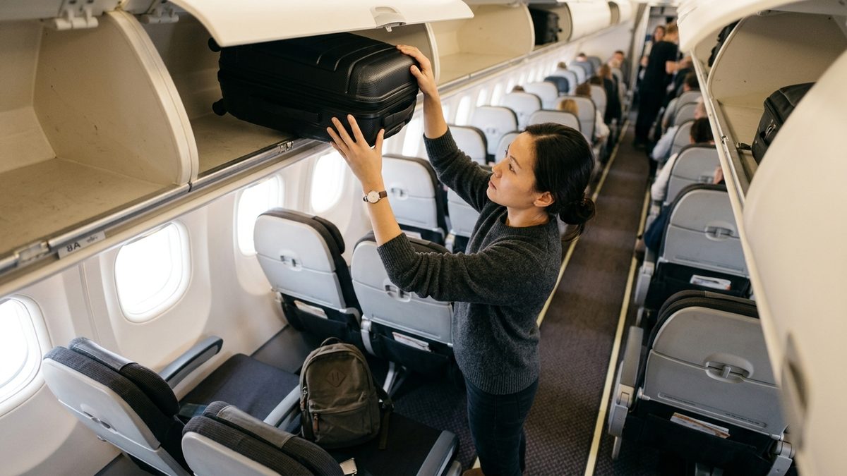 Passenger stowing cabin bag in British Airways overhead locker with underseat backpack visible